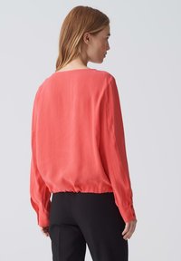 Blusa a maniche lunghe corallo con vestibilità comoda, orlo elastico e polsini abbottonati. Realizzata in tessuto leggero con una consistenza liscia.