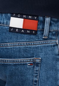 Οπίσθια όψη μπλε τζιν υφάσματος denim με ανάγλυφο λογότυπο Tommy Jeans και μικρή λεπτομέρεια σημαίας κοντά στην πίσω τσέπη, φορεμένα με σκούρο τοπ.