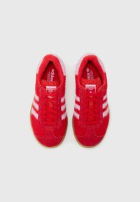 Par de ténis vermelhos da Adidas com riscas brancas e solas cor de borracha, vistos de cima com fundo branco.