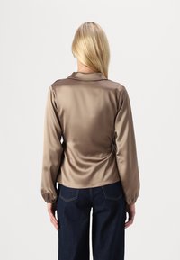 Blusa longa de seda castanha com gola, mangas franzidas e silhueta justa, combinada com jeans de perna larga azul-escuro.