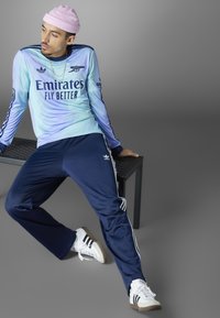 Lysblå lang�ærmet trøje med teksten "Emirates Fly Better", parret med marineblå træningsbukser med hvide striber og Adidas-logo.