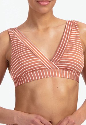 Kobieta ubrana w rdzawobrązowy, biało-paskowany, bezrękawnikowy krótki top z kopertowym dekoltem w serek. Ręce spoczywają w pobliżu talii na jednolitym tle.