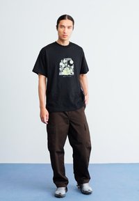 Czarny t-shirt z grafiką w jasnym kolorze; zestawiony z ciemnobrunatnymi spodniami cargo; model w szarych trampkach; jednolite tło.