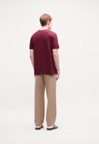 Persoon met een bordeauxrode T-shirt met korte mouwen, beige wijde broek en witte sneakers met zwarte strepen, staand met de rug naar de camera op een witte achtergrond.