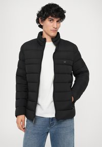 Jachetă puffer neagră cu cusături orizontale, guler înalt, închidere cu fermoar și buzunare. Pur și simplu peste o cămașă albă și asociată cu blugi albaștri.