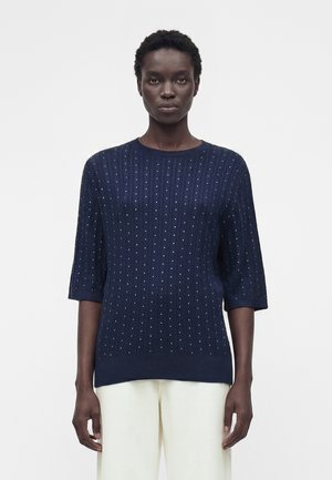 Kvinde med kort krøllet hår iført en navyblå sweater med små hvide prikker og hvide bukser, stående mod en hvid baggrund.