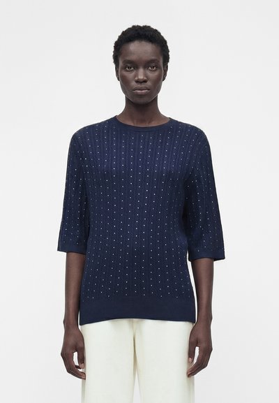 Femme aux cheveux courts et bouclés portant un pull bleu marine à petits pois blancs et un pantalon blanc, debout devant un fond blanc.