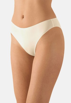 Frau trägt nahtlose beige Bikini-Unterwäsche, zeigt Bauchpartie und obere Oberschenkel vor einfachem hellem Hintergrund.