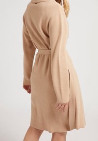 Beiges Strickkleid mit langen Ärmeln, tailliertem Bund, seitlichen Schlitzen und einem Kragen. Weicher Stoff und schlichtes Design.