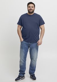 Tamsiai mėlynas medvilninis marškinėliai, laisvas kirpimas, derinami su šviesiai mėlynomis išblukusiomis džinsinėmis kelnėmis. Tamsūs sportbačiai užbaigia aprangą. Paprasta fono.