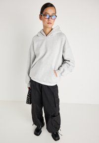 Sivoden kapucar s mehko teksturo, v kombinaciji z ohlapnimi črnimi cargo hlačami z žepi na straneh in črnimi masivnimi čevlji.