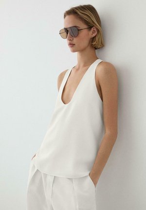 Donna con top bianco senza maniche con scollo a V profondo e pantaloni bianchi, con le mani in tasca e occhiali da aviatore riflettenti, appoggiata a un muro.