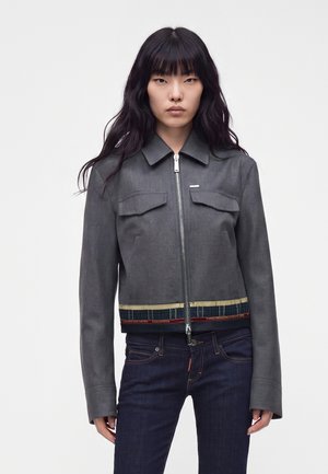 Jeune femme aux longs cheveux noirs portant une veste grise zippée et un jean bleu foncé, debout devant un fond clair uni.