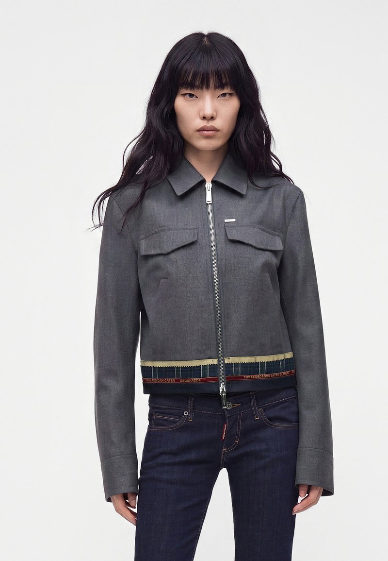 Jeune femme aux longs cheveux noirs portant une veste grise zippée et un jean bleu foncé, debout devant un fond clair uni.