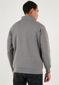 Šedá mikina s vysokým límcem, žebrovanými manžetami a lemem. Hladká textura, minimalistický design bez viditelných log a vzorů.