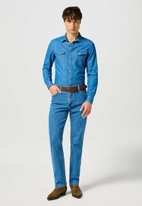 Camisa de denim en azul claro con dos bolsillos en el pecho, costuras en contraste y cuello tipo camisa, combinada con unos jeans de denim azul y un cinturón marrón.