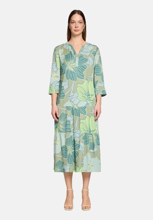 Frau trägt ein Midikleid mit großen grünen und blauen Blumenmustern, Dreiviertelärmeln, V-Ausschnitt und beigen offenen High Heels, steht frontal.