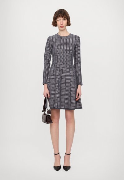 Abito a righe navy con maniche lunghe, gonna svasata, tessuto strutturato e corpetto aderente. La modella tiene in mano una piccola borsa scura e indossa tacchi neri.