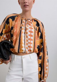 Blusa corta colorida con un cuello atado, mangas largas abullonadas y una mezcla de patrones abstractos en naranja, negro y blanco. Combinada con pantalones blancos.