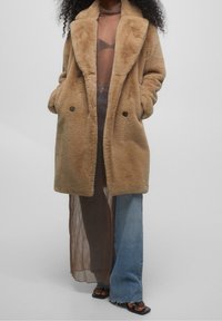 Abrigo de pelo sintético color beige con diseño de doble botonadura, gran cuello con muescas y bolsillos laterales. Llevado sobre un vestido largo transparente y unos pantalones de pierna ancha.