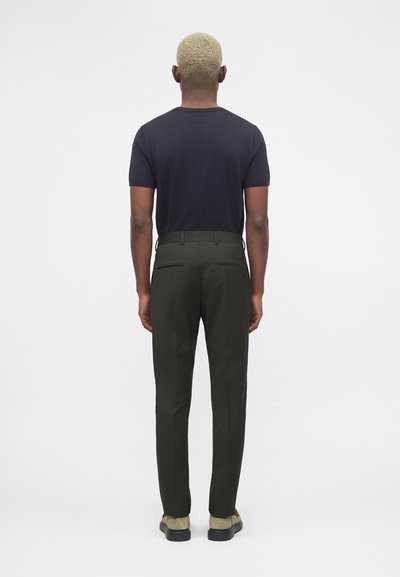 Άνδρας με κοντά ξανθά μαλλιά, που φοράει μαύρο κοντομάνικο πουκάμισο, σκούρα πράσινο παντελόνι και μπεζ παπούτσια, στέκεται γυρισμένος με την πλάτη σε λευκό φόντο.