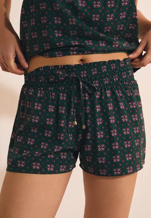 Persoon draagt bijpassende donkergroene korte broek en top met patroon, past de tailleband aan en toont de buik met huidtintcontrast.