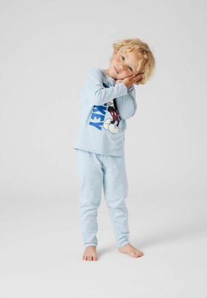Junges Kind mit lockigem, blondem Haar, das hellblaue Mickey-Maus-Pyjamas trägt, den Kopf auf den Händen ruhend, als ob es schläft, barfuß stehend.