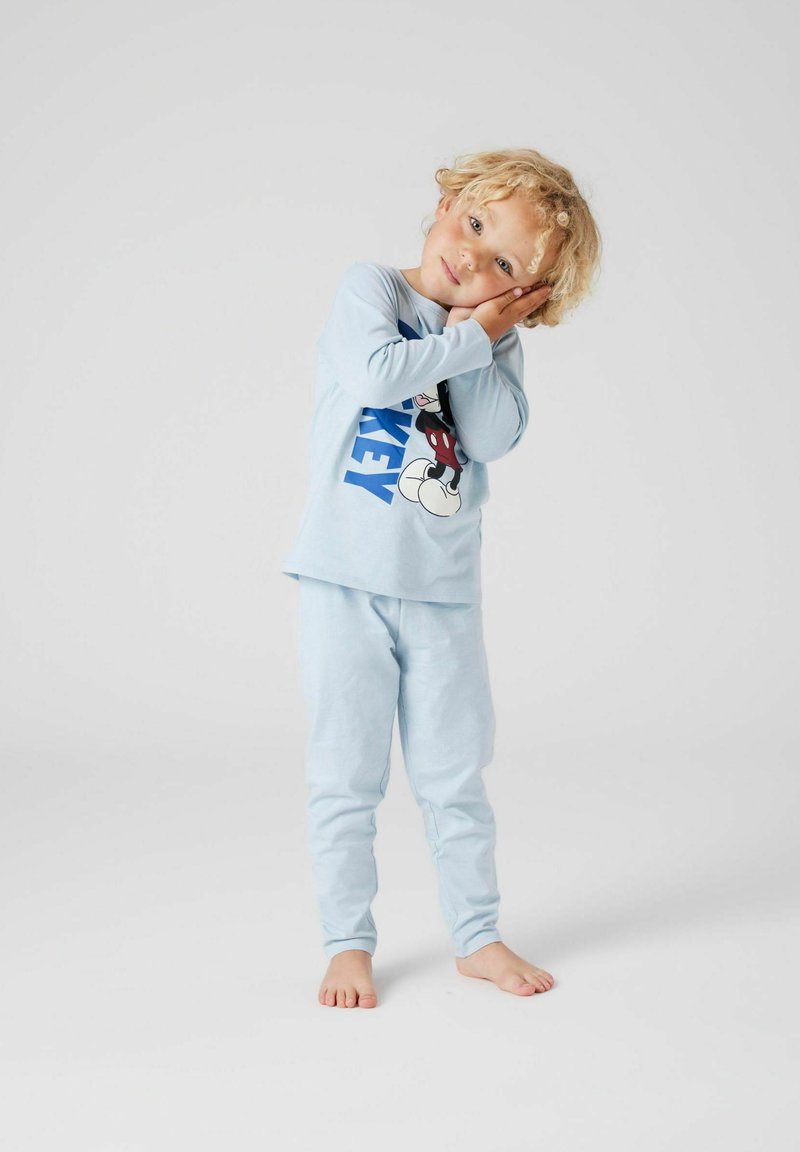 Dziecko o kręconych blond włosach ubrane w jasnoniebieską piżamę z Myszkiem Miki stoi boso, pochylając głowę i opierając ją na rękach, jakby spało.