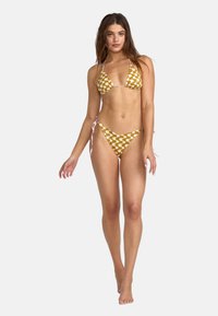 Conjunto de bikini con fondo amarillo mostaza y un patrón ondulado blanco. Incluye una parte superior triangular y una braguita con lazo a los lados, fabricado en un tejido ligero.