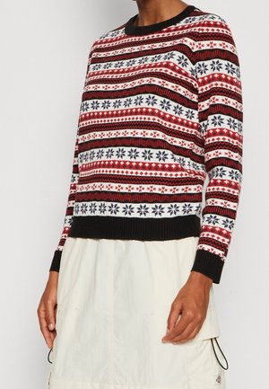 Henkilö, joka pukeutunut punaiseen, valkoiseen ja mustaan kuvioituun neuleeseen, jossa on lumihiutaledesignit, ja valkoiseen hameeseen, jossa on taskudetalji.