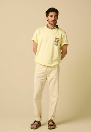 Homme portant un t-shirt jaune pâle avec un motif coquillage, pantalon crème, sandales marron et foulard à motifs autour du cou, debout les mains dans le dos.