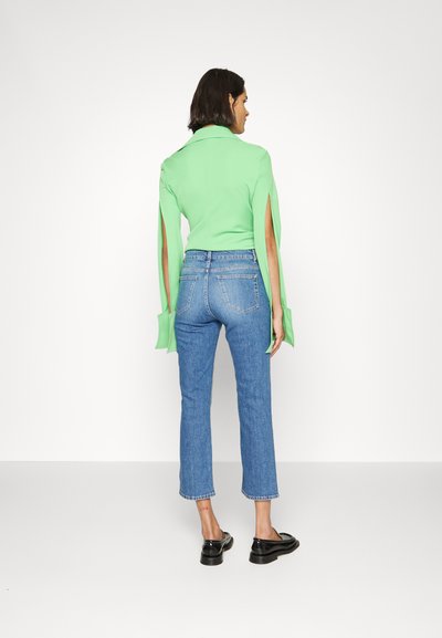 Grünes, langärmliges Oberteil mit seitlichen Schlitzen, kombiniert mit blauen, geraden Jeans und schwarzen Slippers. Rückansicht, minimalistisches Design.