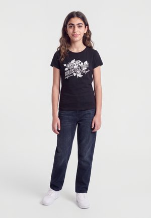 Teenage pige står, iført en sort t-shirt med grafik, mørke jeans og hvide sneakers mod en enkel lys baggrund.