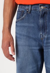 Jean en denim bleu avec une coupe regular, présentant des coutures contrastées, cinq poches et une fermeture à bouton en métal au niveau de la taille.