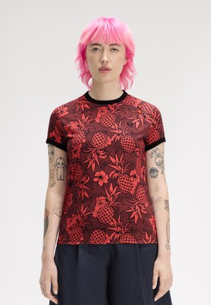 Person mit leuchtend pinkem Haar und Tätowierungen trägt ein rotes Hemd mit Ananas-Muster und schwarzem Rand sowie dunkle Hosen vor schlichtem Hintergrund.