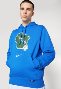 Modrá mikina s kapucňou vyrobená z jemného materiálu, s tvarom Wisconsinu v zelenej farbe a textom "BUCKS" spolu s grafickým znázornením basketbalu. Na prednej strane je biele logo Nike.