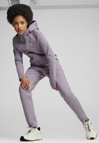 Sudadera con cremallera morada y joggers a juego, hechos de material suave, con un logo de Puma en negro, combinados con zapatillas blancas con acentos negros.