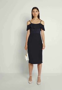 Vestido preto sem mangas com decote pregueado, silhueta ajustada, até ao joelho, combinado com uma pequena mala branca e sapatos de salto beje.
