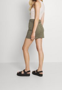Mini-jupe vert olive au design près du corps, associée à des sandales à plateforme noires. Tissu lisse avec un léger éclat et des détails de surpiqûre subtils.