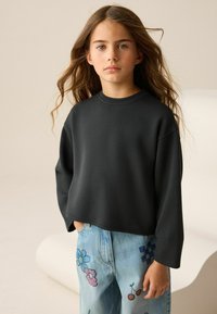 Ein Mädchen mit langen Haaren, das einen schwarzen übergroßen Sweatshirt und eine blaue Jeans trägt, die mit bunten Blumen-, Kirsch- und karierten Aufnähern verziert sind.