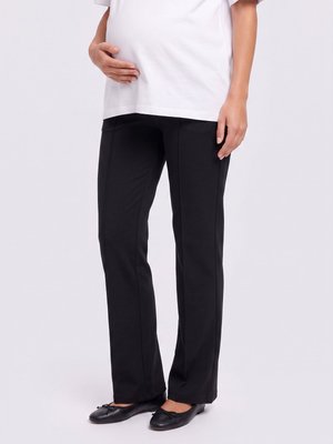 Persona embarazada que lleva pantalones premamá negros, camiseta blanca y zapatillas negras, con una mano sobre la barriga y la otra relajada a un lado.