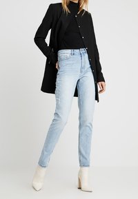Blazer negro de manga larga, tejido texturizado, con cierre de botones, combinado con jeans desgastados de cintura alta en azul claro y botas de tacón alto en color crema.