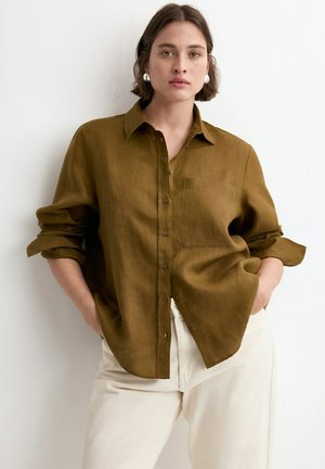 Mujer vestida con camisa verde oliva abotonada con mangas remangadas y pantalones blancos, de pie contra una pared blanca lisa, con las manos en los bolsillos.