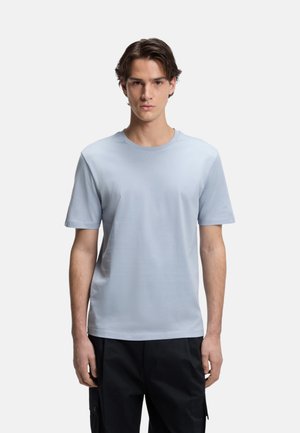 Młody mężczyzna z krótkimi brązowymi włosami, ubrany w jasnoniebieską koszulkę z krótkim rękawem i czarne spodnie, stojący na jednolitym białym tle.