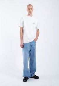 Camiseta blanca de algodón con mangas cortas y un pequeño logo negro en el pecho, combinada con unos vaqueros de mezclilla de corte holgado en azul claro y zapatillas negras.