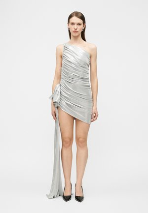Vrouw draagt een zilverkleurige mini-jurk met een enkele schouder, geplooid, met een grote strik aan de zijkant en een lange stoffen staart, gecombineerd met zwarte puntige hakken.