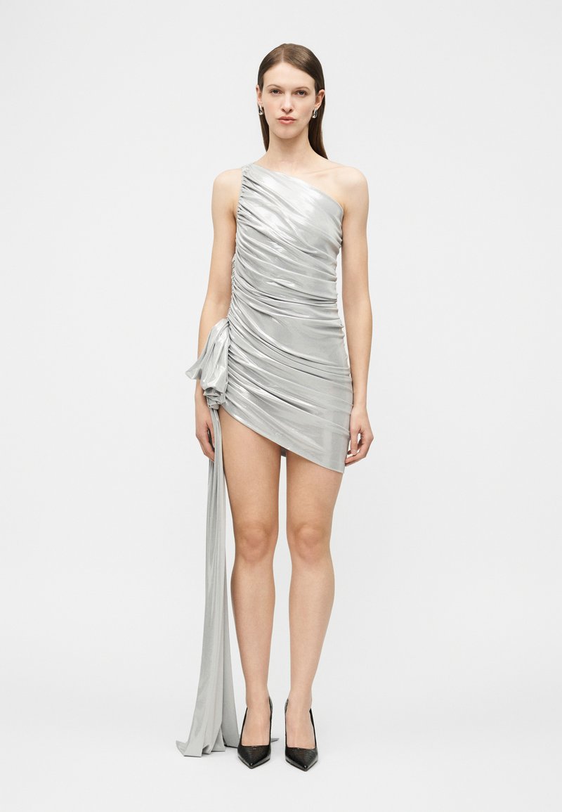 Γυναίκα που φοράει ασημί μίνι φόρεμα με έναν ώμο, ρυτιδιασμένο, με μεγάλο πλευρικό φιόγκο και μακριά ουρά υφάσματος, συνδυασμένο με μαύρες μυτερές γόβες.