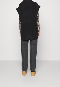 Svart stickad ärmlös tröja med ribbad fåll, bärs över en prickig topp, kombinerad med mörkgrå lösa jeans och ljusbruna platåskor.