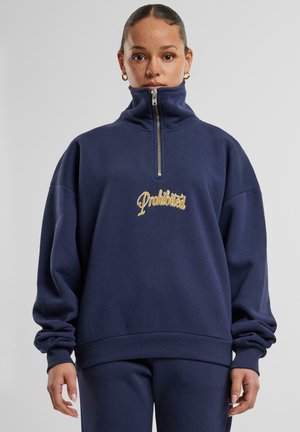 Mørkeblå genser med høy krage, frontglidelås, og brodert "Forbudt"-logo i gull. Har avslappet passform og ribbestrikkede mansjetter.
