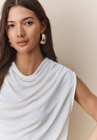 White draped top with soft, flowing fabric; features a high neckline and sleeveless design. Model wears large, ornate gold hoop earrings.